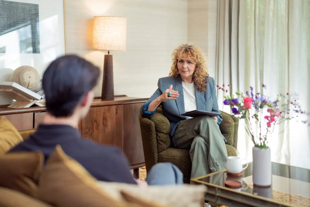 Vrouw in een blauwe blazer zit in een groene fauteuil en gebaart tijdens een gesprek met een man die op de voorgrond zit. Ze houdt een tablet op haar schoot. Naast haar staat een tafel met een witte kop en een vaas met kleurrijke bloemen. De ruimte heeft warme houten tinten, zachte gordijnen en veel natuurlijk licht.