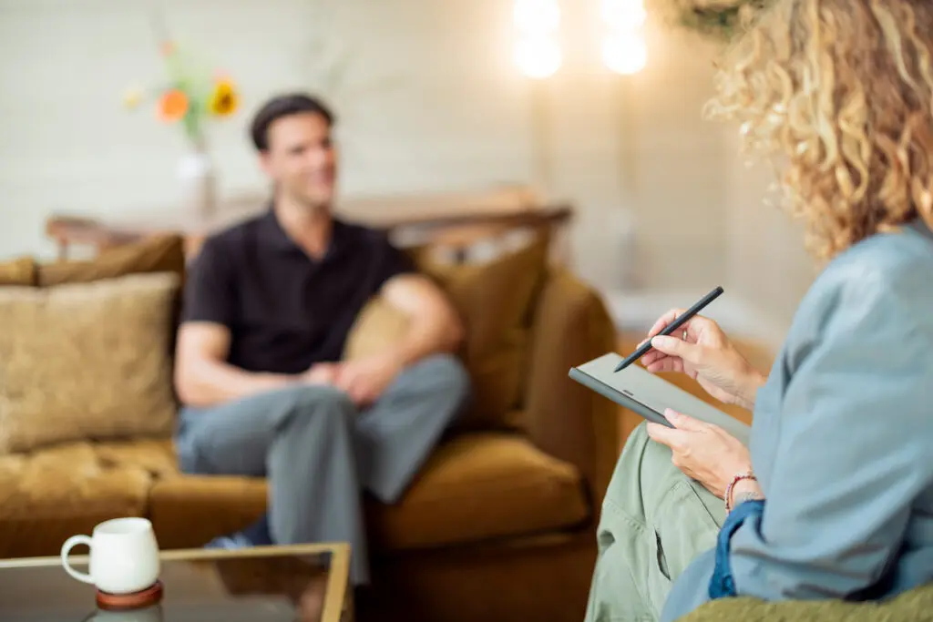 Vrouw in een blauwe blouse zit met een notitieboek op schoot en maakt aantekeningen tijdens een gesprek met een man die tegenover haar op een bruine bank zit. Op de voorgrond staat een witte kop op een houten tafel. De ruimte heeft warme bruine tinten, zachte verlichting en een huiselijke sfeer.