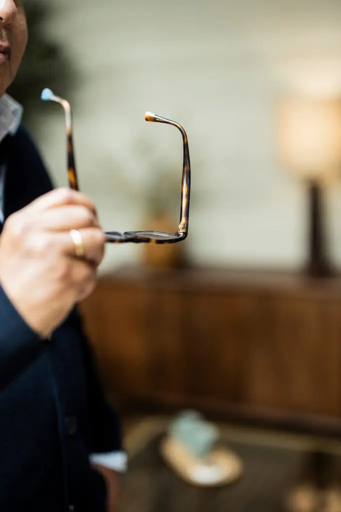 Persoon in donkere kleding houdt een donkere bril in de hand. De bril staat centraal in beeld en is scherp afgebeeld, terwijl de persoon en de warme, sfeervolle ruimte op de achtergrond onscherp zijn.