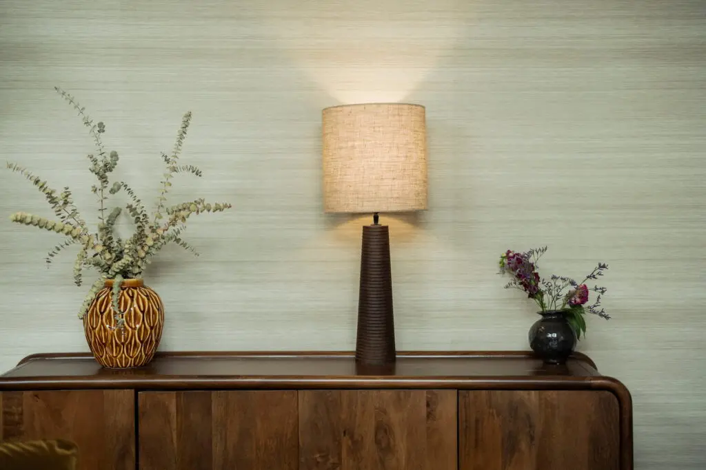 Houten dressoir tegen een lichtgroene wand, met in het midden een brandende tafellamp en aan weerszijden twee decoratieve vazen met groene en paarse bloemen. De warme houttinten en zachte verlichting zorgen voor een rustige, huiselijke sfeer.