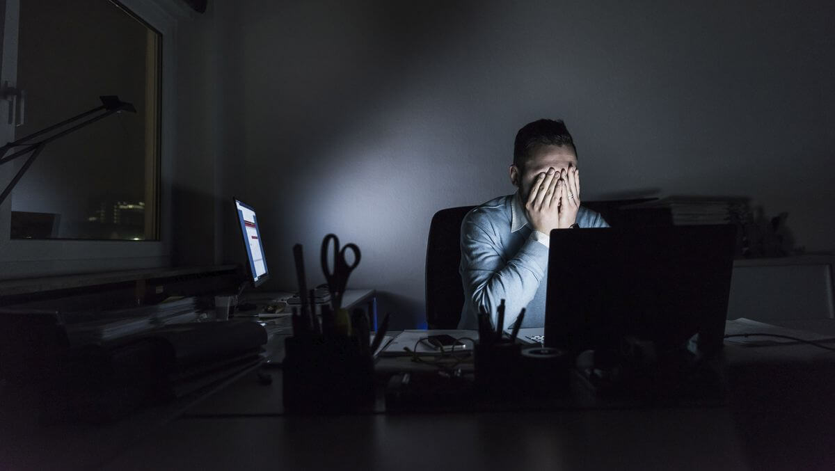 Man zit ’s avonds achter een laptop met zijn handen voor zijn gezicht, zichtbaar vermoeid of gestrest