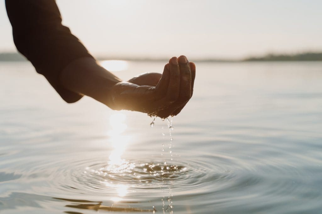 Hand schept water uit een rustig oppervlak terwijl druppels terugvallen in het water bij zonlicht