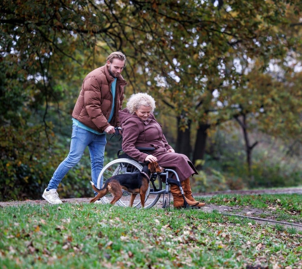 Man helpt vrouw in rolstoel tijdens wandeling in park, symbool voor zorg, ondersteuning en welzijn