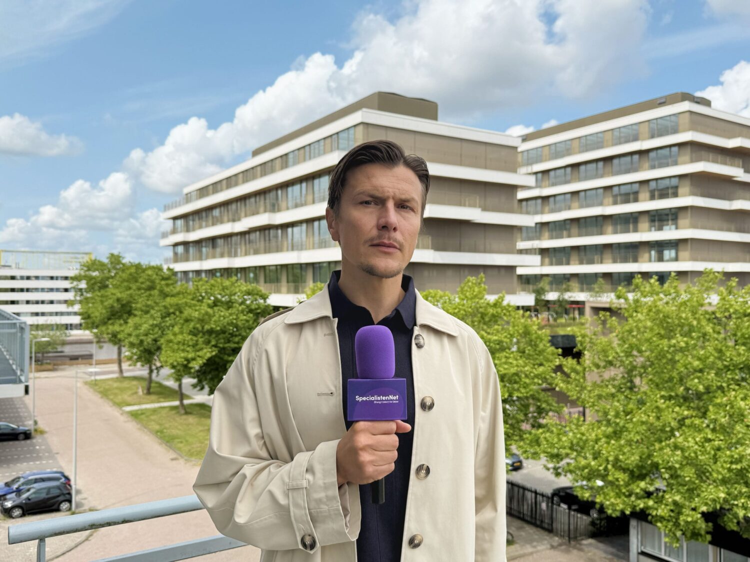 Man met paarse microfoon staat buiten voor modern kantoorgebouw en doet verslag