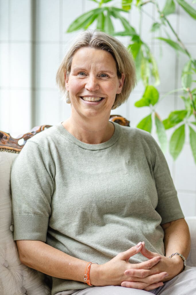 Vrouw met groen shirt en vriendelijke uitstraling zit op stoel en glimlacht