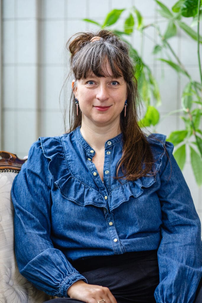 Vrouw met blauwe blouse en vriendelijke uitstraling zit op stoel