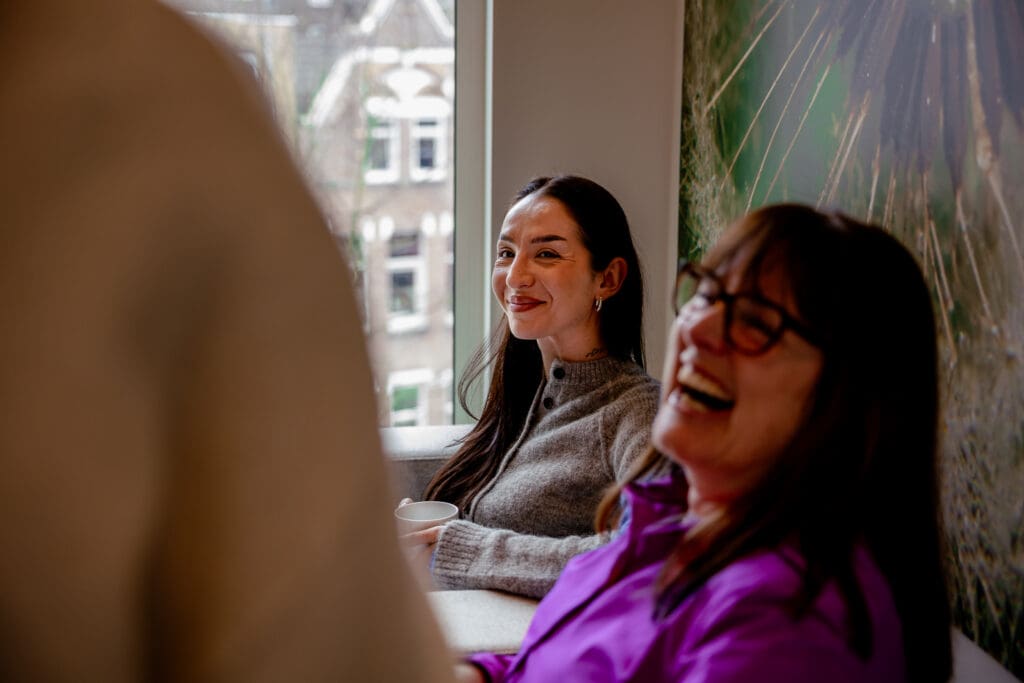 Twee collega’s zitten lachend met elkaar in gesprek in een lichte kantoorruimte. Een van hen houdt een koffiekop vast, terwijl op de voorgrond een derde persoon onscherp zichtbaar is. De afbeelding straalt een informele en collegiale werksfeer uit.