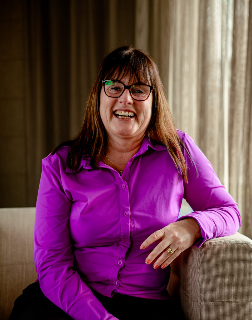 Professioneel portret van een glimlachende medewerker met een paarse blouse en bril, zittend op een fauteuil in een rustige kantooromgeving. De medewerker kijkt recht in de camera en leunt ontspannen op de armleuning.