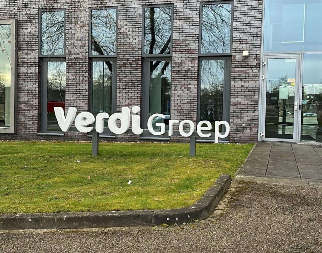 Grote witte VerdiGroep-belettering op een gazon voor een modern kantoorgebouw met grijsbruine bakstenen, hoge ramen en een glazen entree.