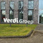 Grote witte VerdiGroep-belettering op een gazon voor een modern kantoorgebouw met grijsbruine bakstenen, hoge ramen en een glazen entree.