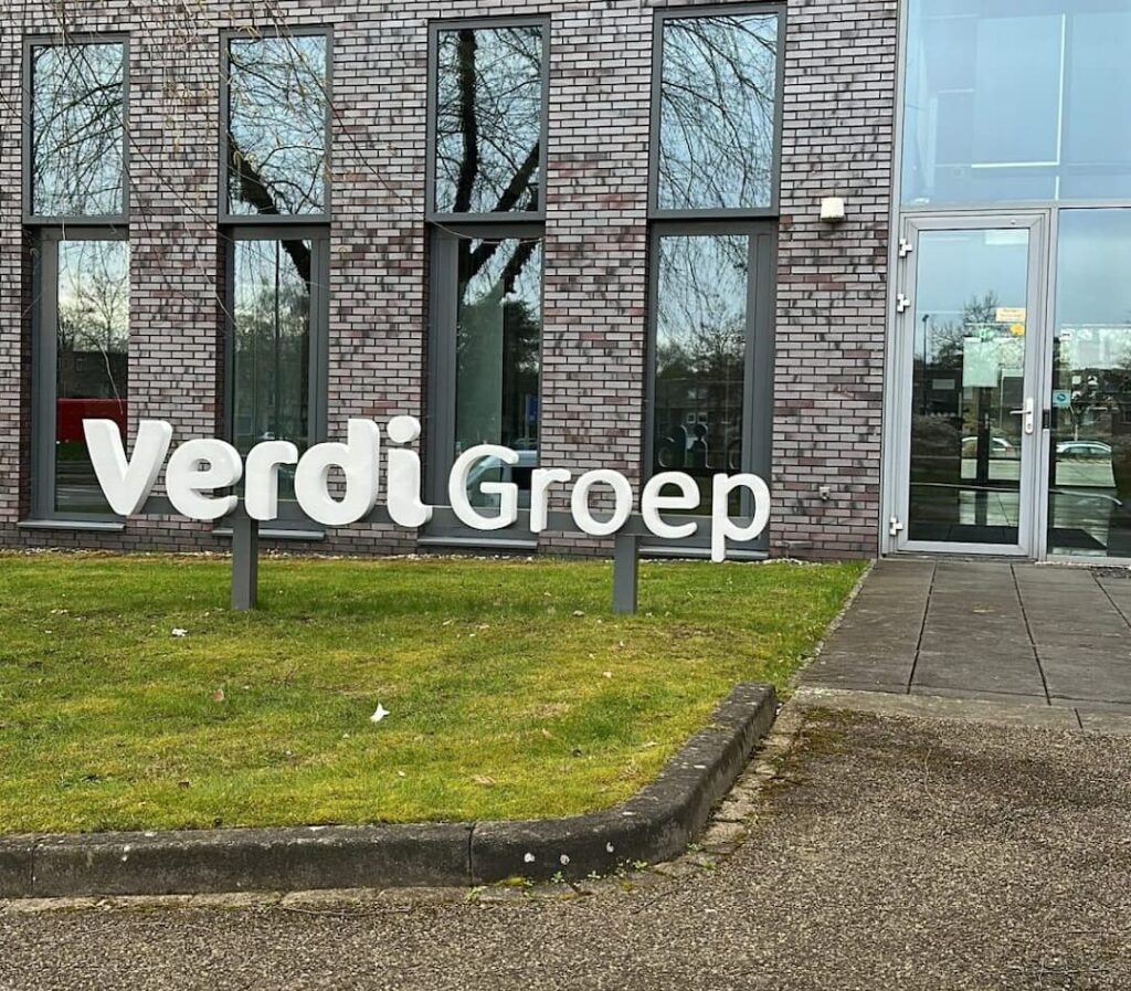 Grote witte VerdiGroep-belettering op een gazon voor een modern kantoorgebouw met grijsbruine bakstenen, hoge ramen en een glazen entree.