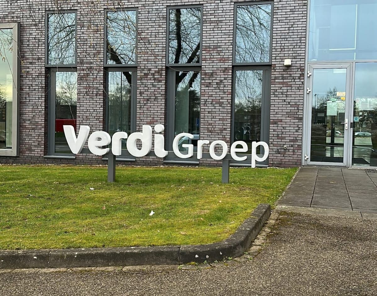 Grote witte VerdiGroep-belettering op een gazon voor een modern kantoorgebouw met grijsbruine bakstenen, hoge ramen en een glazen entree.