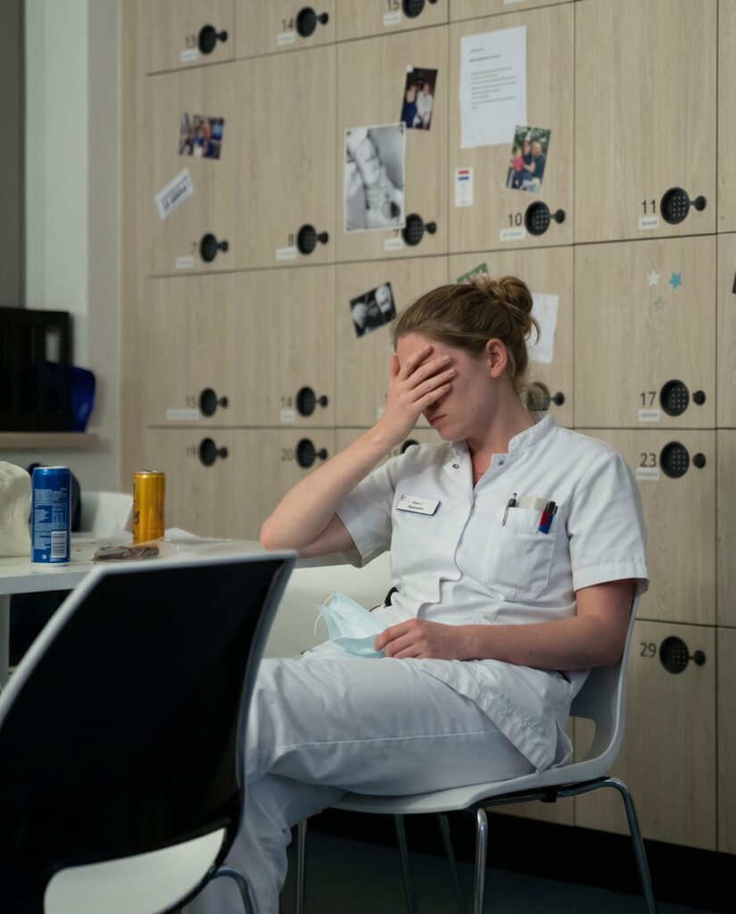 Vermoeide zorgmedewerker in wit uniform zit tijdens een pauze met hand voor het gezicht in een personeelsruimte met lockers
