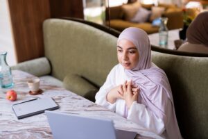 Vrouw met een lichtpaarse hijab zit aan een marmeren tafel met haar handen tegen haar borst, naast een laptop, notitieboek, kop koffie en appel. Achter haar is een groene bank zichtbaar.
