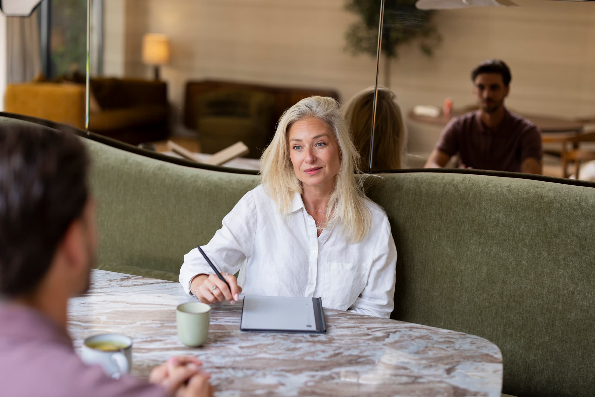 Blonde coach zit aan een tafel met een notitieblok en pen en luistert aandachtig naar een cliënt tegenover haar.