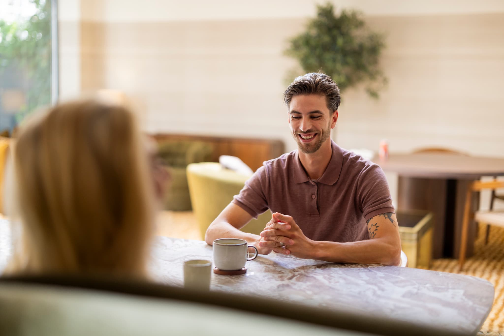 Man in een roze poloshirt zit lachend aan tafel met zijn handen gevouwen, terwijl hij met een gesprekspartner praat in een sfeervolle ruimte.