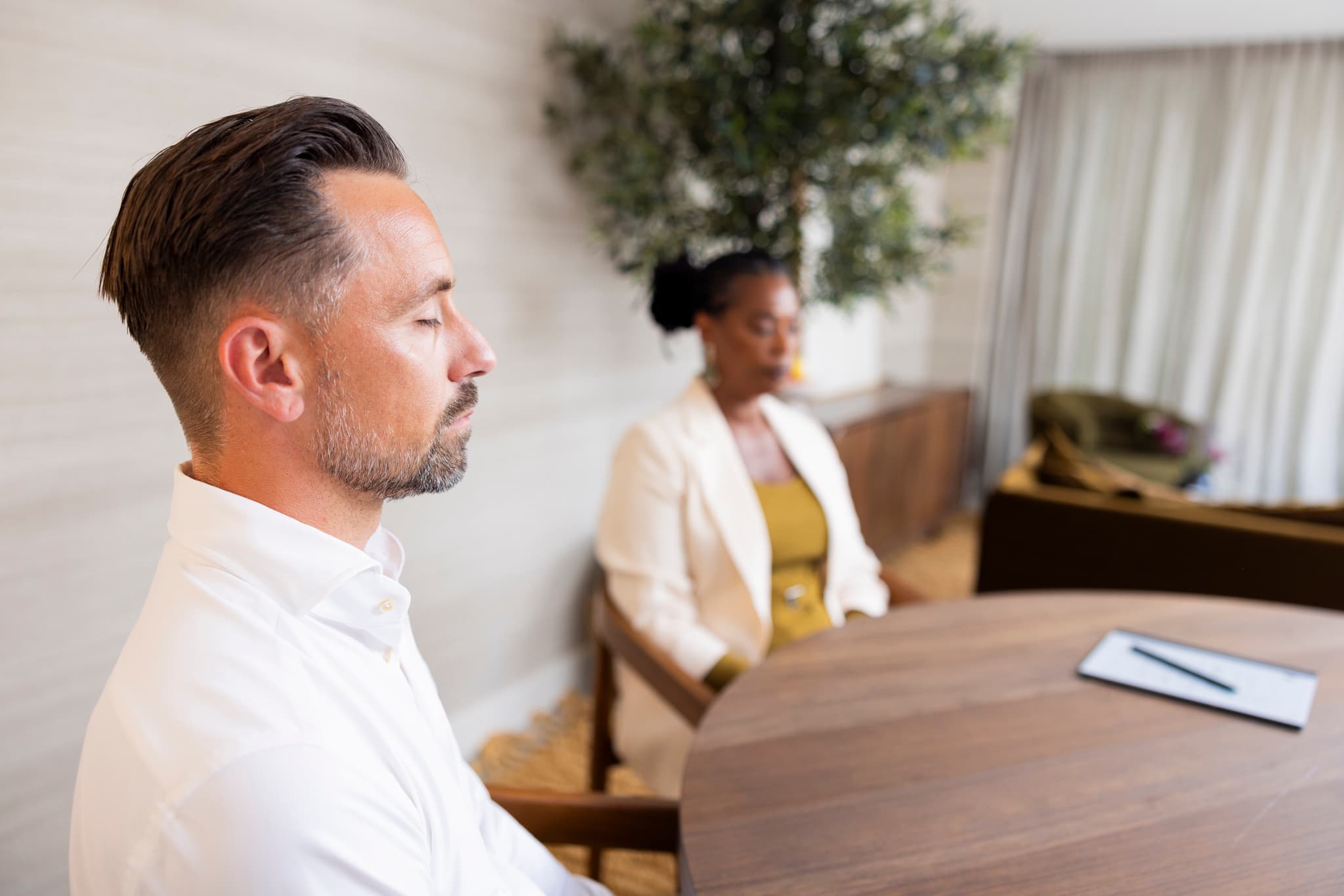 Een man en een vrouw zitten tegenover elkaar aan een ronde houten tafel in een rustige, modern ingerichte ruimte. Op tafel ligt een notitieblok met pen. De man is op de voorgrond scherp in beeld, terwijl de vrouw op de achtergrond zit.