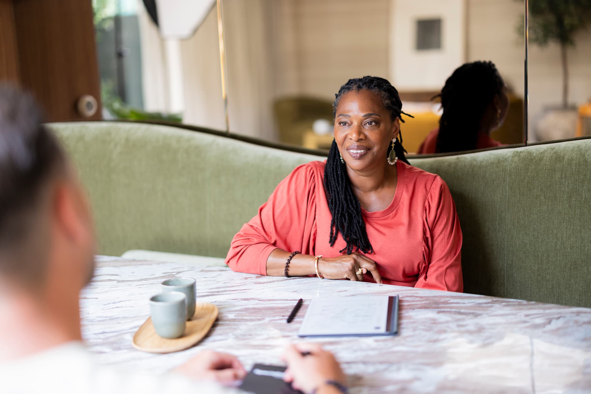 Een vrouw in een koraalrode top zit aan een tafel in een groene bank en kijkt naar haar gesprekspartner. Op tafel staan twee kopjes, een notitieblok en een pen. De gesprekspartner is op de voorgrond onscherp zichtbaar.