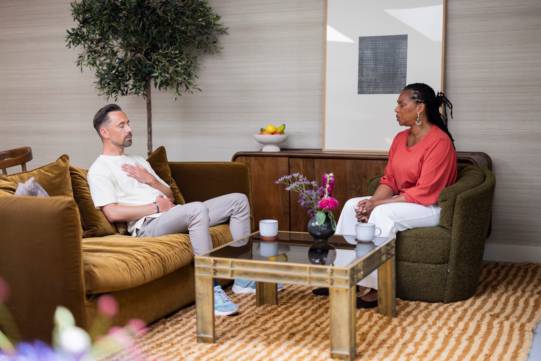 Een man en een vrouw zitten tegenover elkaar in een sfeervolle woonkamer en zijn met elkaar in gesprek. De man zit op een bruine bank en de vrouw in een groene fauteuil. Tussen hen staat een glazen salontafel met bloemen en twee kopjes.