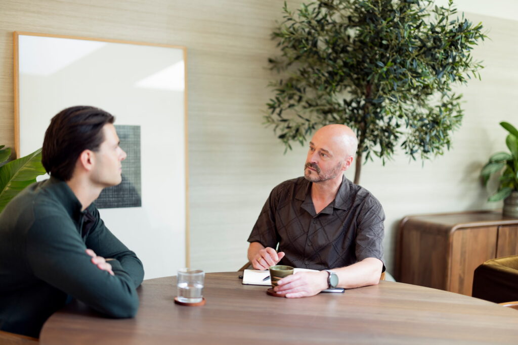 Twee mannen zitten tegenover elkaar aan een houten tafel en voeren een aandachtig gesprek. De man rechts houdt een kopje vast, met een notitieboek en pen voor zich. De man links luistert met zijn armen over elkaar. Op de achtergrond staan een grote plant en een ingelijste afbeelding in een lichte, warme ruimte.