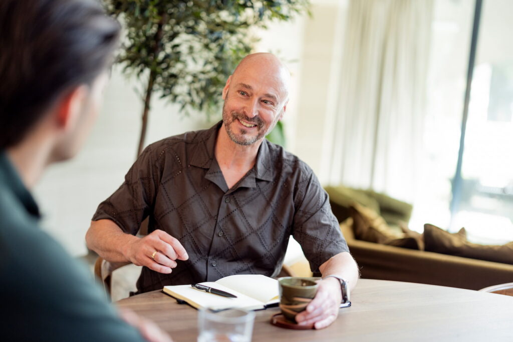 Man in een donker overhemd zit aan een houten tafel en kijkt glimlachend naar een persoon op de voorgrond. Voor hem liggen een open notitieboek met een pen en een kopje. De lichte ruimte heeft warme tinten, grote ramen en een groene plant op de achtergrond.