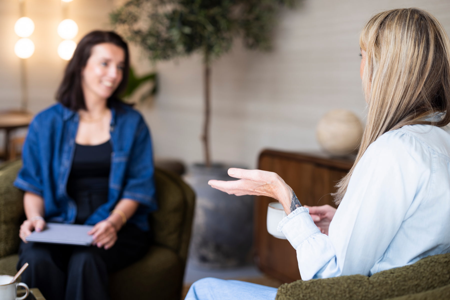 Twee vrouwen zitten tegenover elkaar in comfortabele groene fauteuils en zijn met elkaar in gesprek. De vrouw op de voorgrond houdt een witte kop vast en maakt een handgebaar, terwijl de andere vrouw met een notitieboek op haar schoot aandachtig luistert. De ruimte heeft warme, neutrale tinten en zachte verlichting.