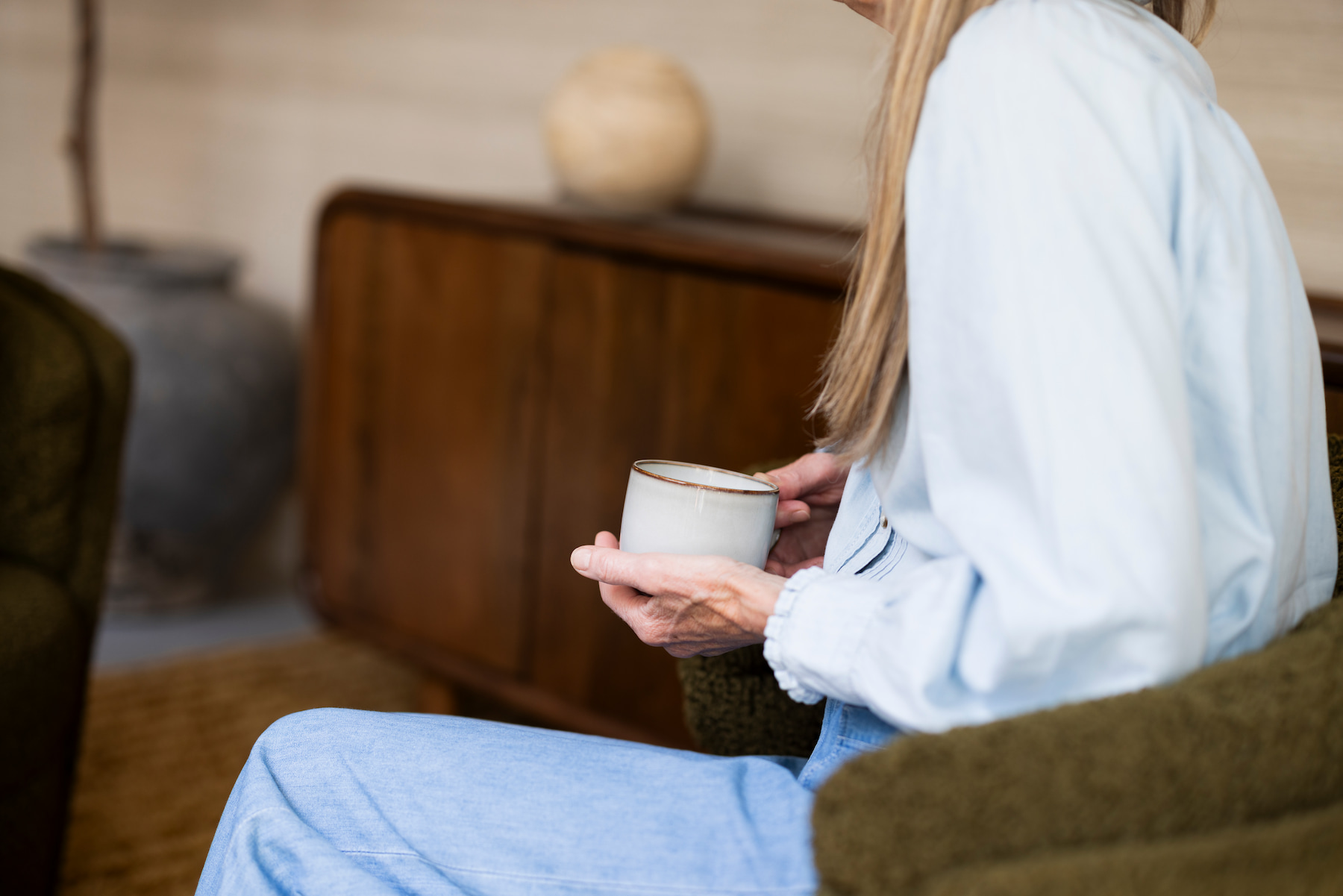 Vrouw in een lichtblauwe blouse en blauwe broek zit in een groene fauteuil en houdt een witte kop koffie met beide handen vast. Op de achtergrond staat een houten kast in een warme, huiselijke ruimte.