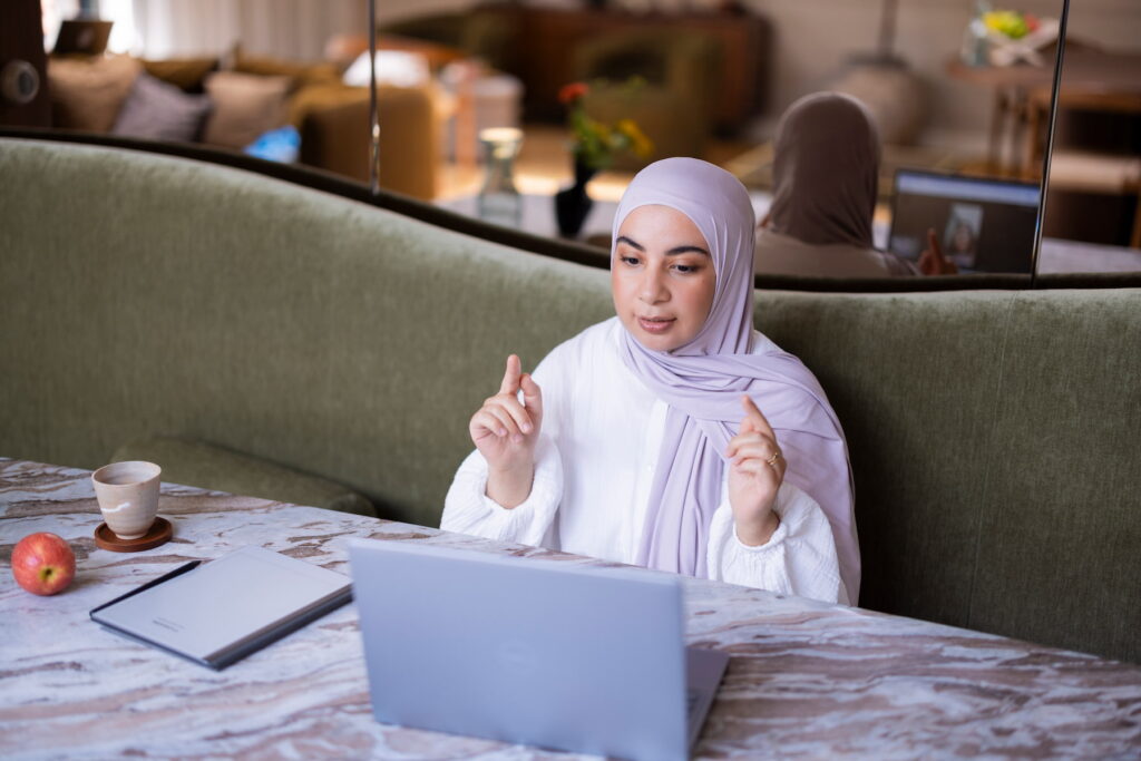 Vrouw met een lichtpaarse hoofddoek zit aan een tafel achter een laptop en maakt met beide handen een gebaar tijdens een gesprek. Op tafel staan een kop koffie en een appel en ligt een notitieboek. Op de achtergrond zijn een groene bank en een spiegel zichtbaar.