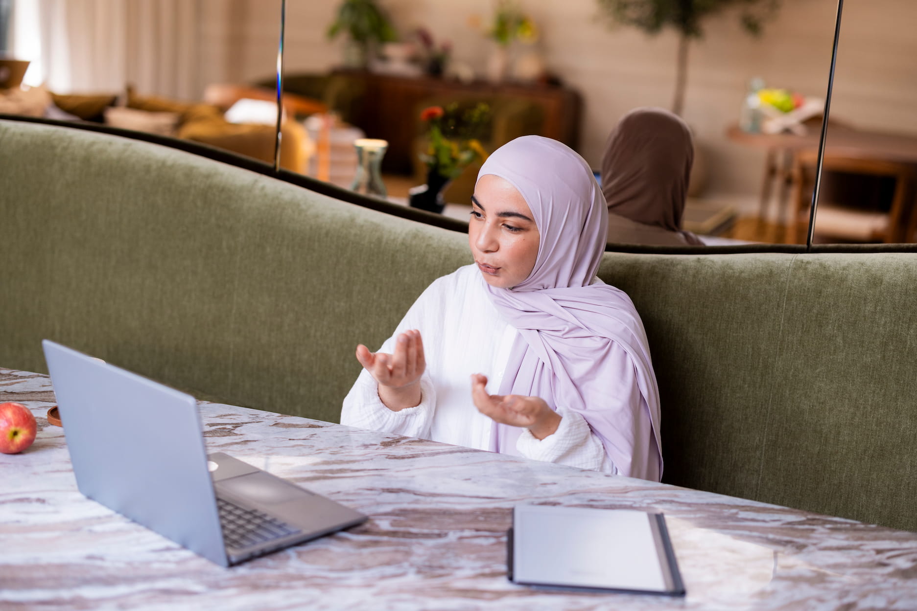 Vrouw met een lichtpaarse hoofddoek zit aan een tafel met een open laptop en maakt een gebaar met haar handen. Voor haar ligt een notitieboek. Op de achtergrond zijn een groene bank, een spiegel en een sfeervol ingerichte ruimte zichtbaar.