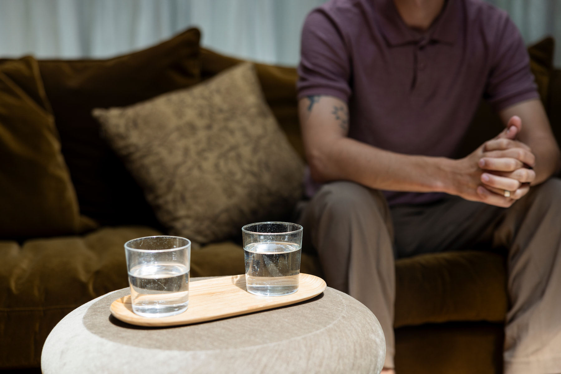 Man in een roze shirt zit op een bruine bank met zijn handen samengevouwen. Op de voorgrond staan twee glazen water op een houten dienblad op een ronde tafel. Bruine kussens en lichte gordijnen zorgen voor een warme, rustige sfeer.