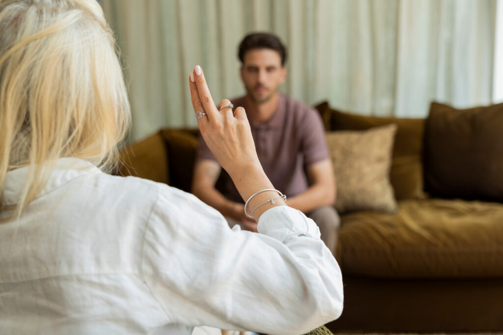 Persoon in witte kleding steekt tijdens een gesprek een hand op, terwijl een man op de achtergrond op een bruine bank zit en luistert. De ruimte heeft lichte gordijnen, warme bruine tinten en kussens op de bank.