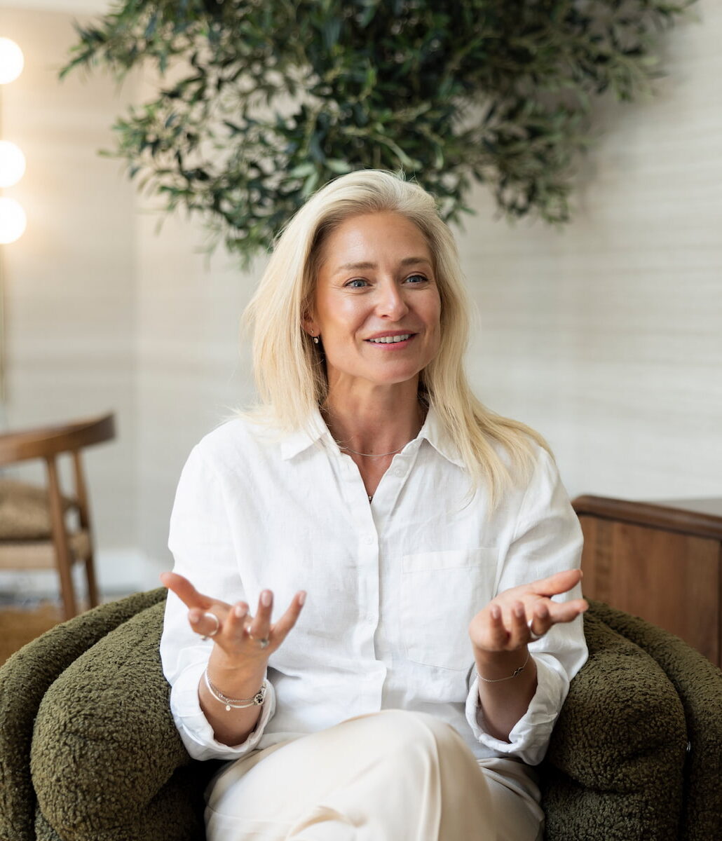 Vrouw in witte kleding zit in een groene fauteuil en praat met open handen tijdens een gesprek. Op de voorgrond is de andere persoon van achteren zichtbaar. De rustige ruimte heeft warme verlichting, een houten interieur en een grote groene plant.