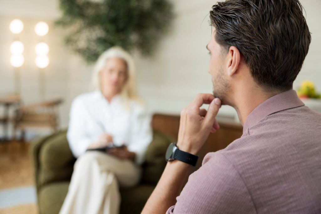 Man in een roze shirt zit op de voorgrond en raakt nadenkend zijn kin aan tijdens een gesprek. Op de achtergrond zit een vrouw in witte kleding met een tablet in een groene fauteuil. De warme, rustige ruimte heeft planten en sfeervolle verlichting.