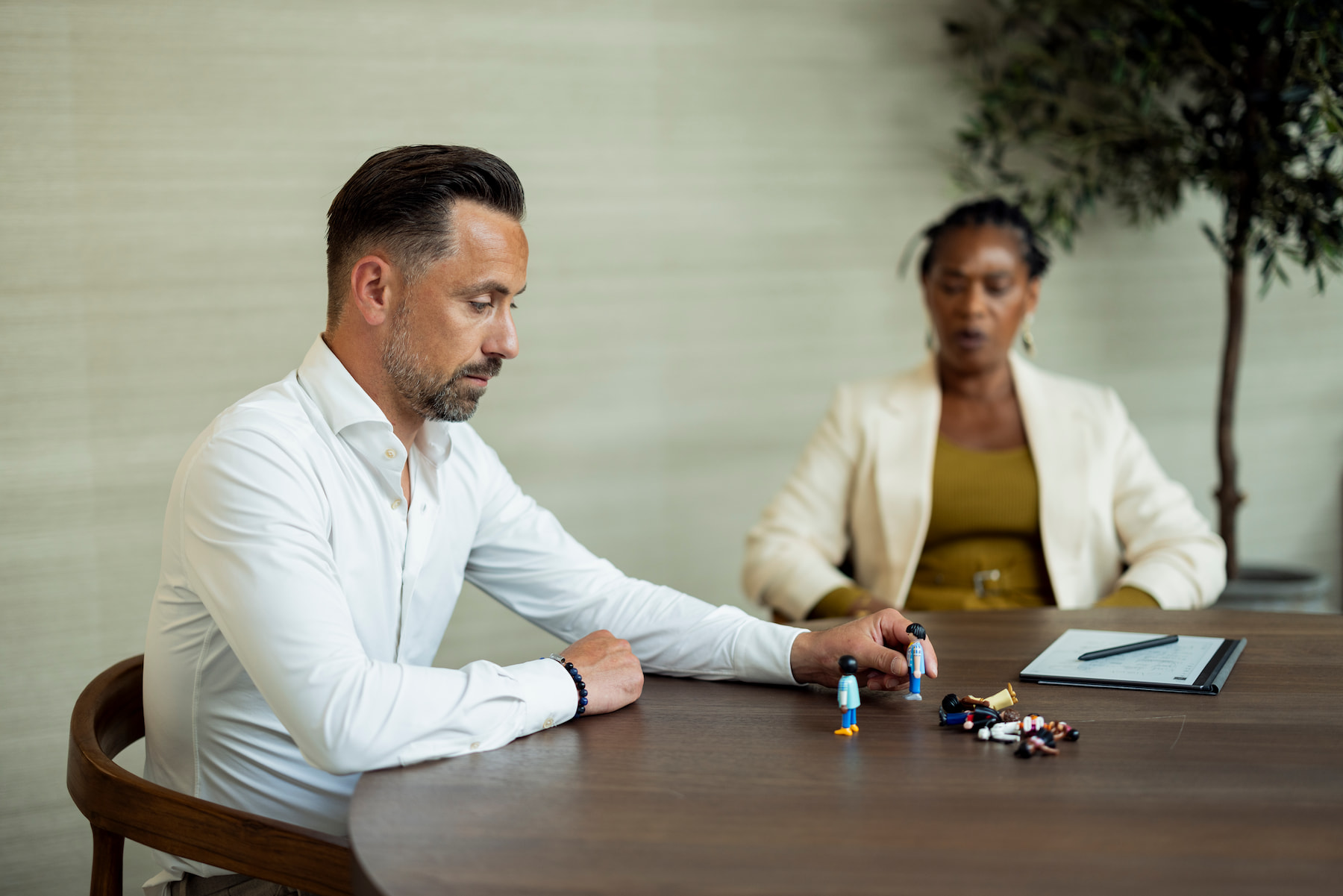 Een man zit aan een tafel en plaatst kleine figuren terwijl een vrouw tegenover hem toekijkt. Op tafel liggen meerdere figuren en een notitieblok met pen. De rustige ruimte heeft een warme, professionele uitstraling.