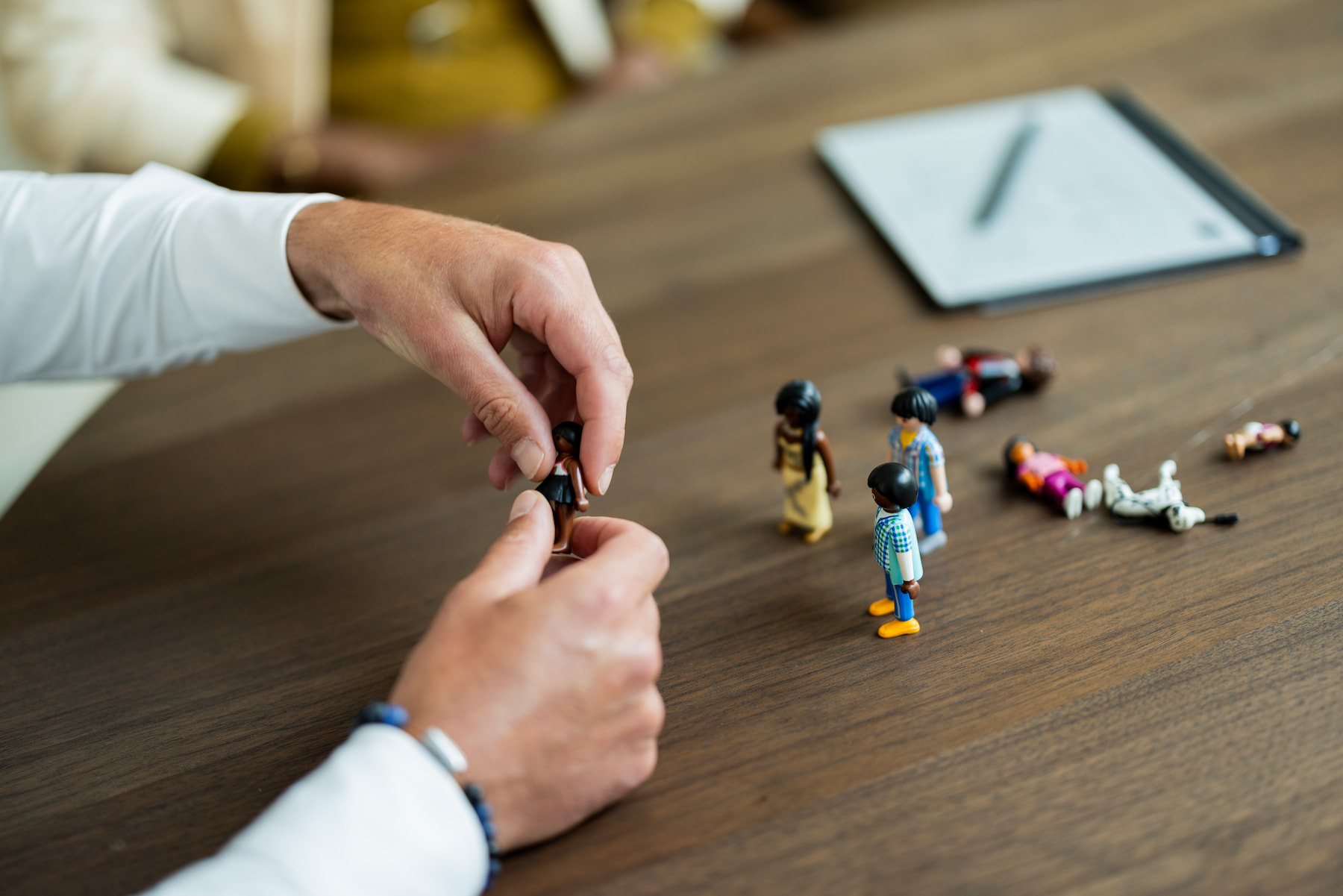 Een persoon verplaatst kleine speelgoedfiguren op een houten tafel. Meerdere figuren staan verspreid op tafel, terwijl andere op hun zij liggen. Op de achtergrond ligt een notitieblok met pen.