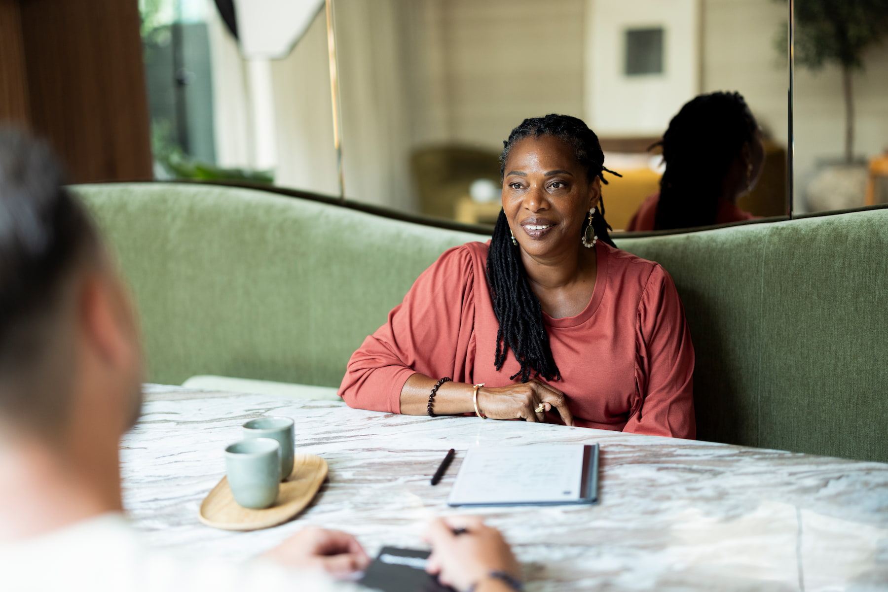 Een vrouw zit glimlachend aan een tafel en kijkt naar haar gesprekspartner op de voorgrond. Op tafel liggen een notitieblok en pen en staan twee kopjes. Een spiegel op de achtergrond weerspiegelt de warme, huiselijke ruimte en de aanwezige gesprekspartners.