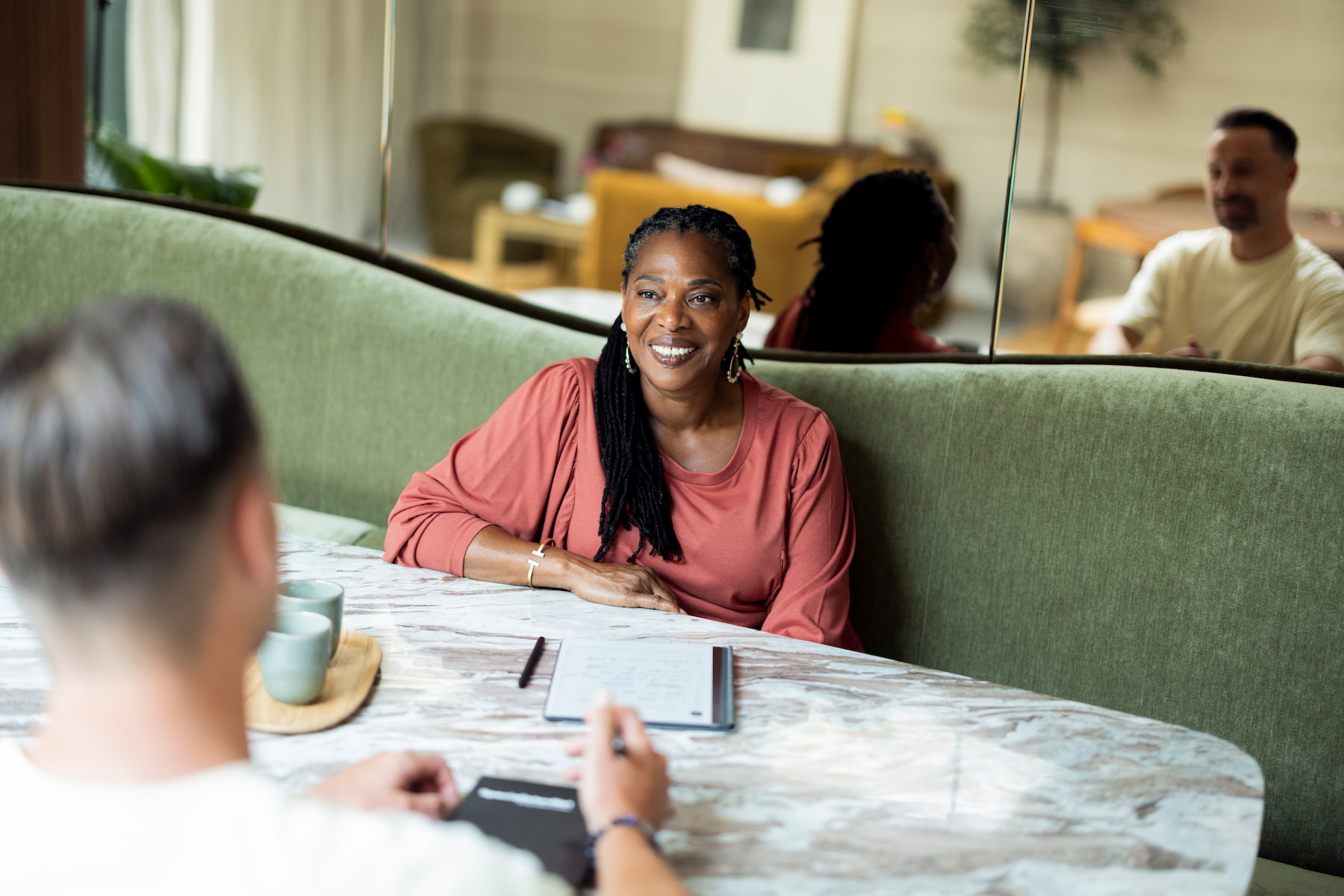 Een vrouw zit glimlachend aan een tafel en kijkt naar haar gesprekspartner op de voorgrond. Op tafel liggen een notitieblok en pen en staan twee kopjes. Een spiegel op de achtergrond weerspiegelt de warme, huiselijke ruimte en de aanwezige gesprekspartners.