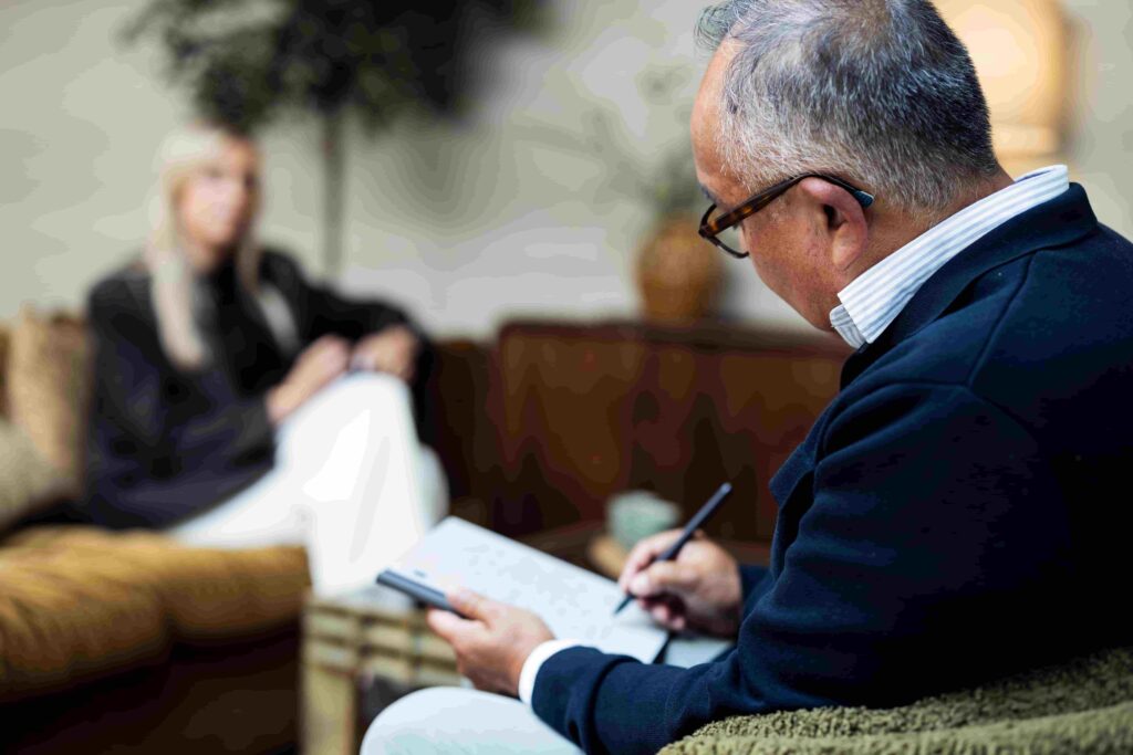 Een oudere man met een bril maakt aantekeningen op een notitieblok terwijl hij tegenover een vrouw zit. De vrouw is op de achtergrond onscherp zichtbaar in een warme, huiselijke zithoek.