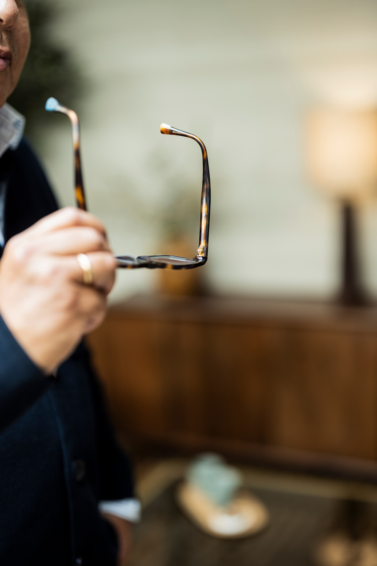 Persoon in donkere kleding houdt een donkere bril in de hand. De bril staat centraal in beeld en is scherp afgebeeld, terwijl de persoon en de warme, sfeervolle ruimte op de achtergrond onscherp zijn.