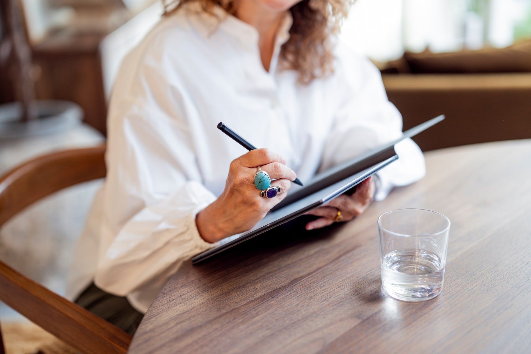 Vrouw in een witte blouse schrijft met een zwarte pen in een donkere map terwijl ze aan een houten tafel zit. Ze draagt opvallende ringen met blauwe stenen. Naast haar staat een glas water. De achtergrond is licht en sfeervol met veel natuurlijk licht.