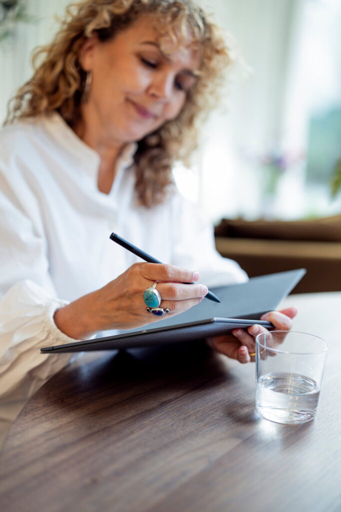 Vrouw met krullend haar en een wit overhemd houdt een open notitieboek vast en schrijft met een pen. Op de houten tafel staat een glas water. De achtergrond is licht en onscherp, met veel natuurlijk licht.