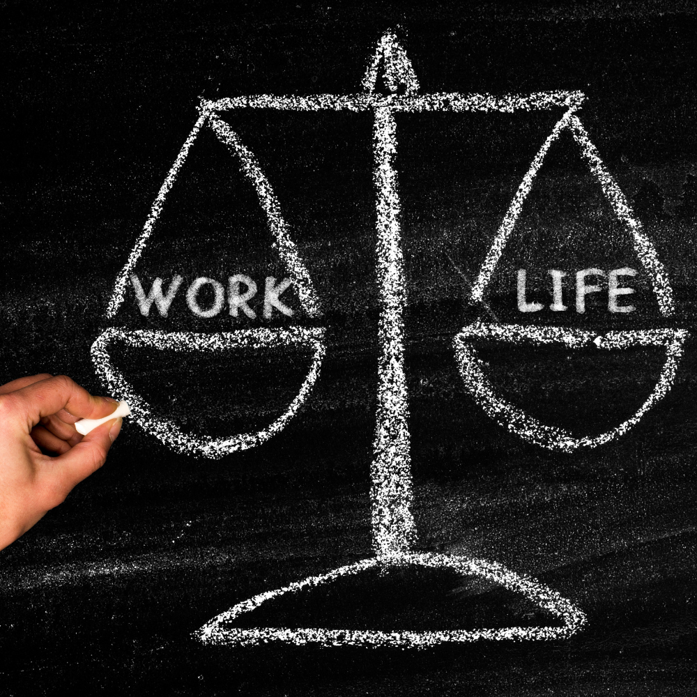 Een witte krijttekening van een weegschaal op een zwart schoolbord, met het woord ‘WORK’ op de linker schaal en ‘LIFE’ op de rechter schaal. Links onderaan tekent een hand met wit krijt aan de weegschaal. De afbeelding symboliseert de balans tussen werk en privéleven.
