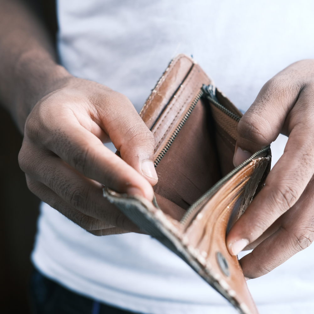 Close-up van twee handen die een lege, bruine leren portemonnee met ritssluiting openhouden. De lege vakken zijn duidelijk zichtbaar. De persoon draagt een wit shirt. De afbeelding illustreert geldgebrek, financiële zorgen of een lege portemonnee.